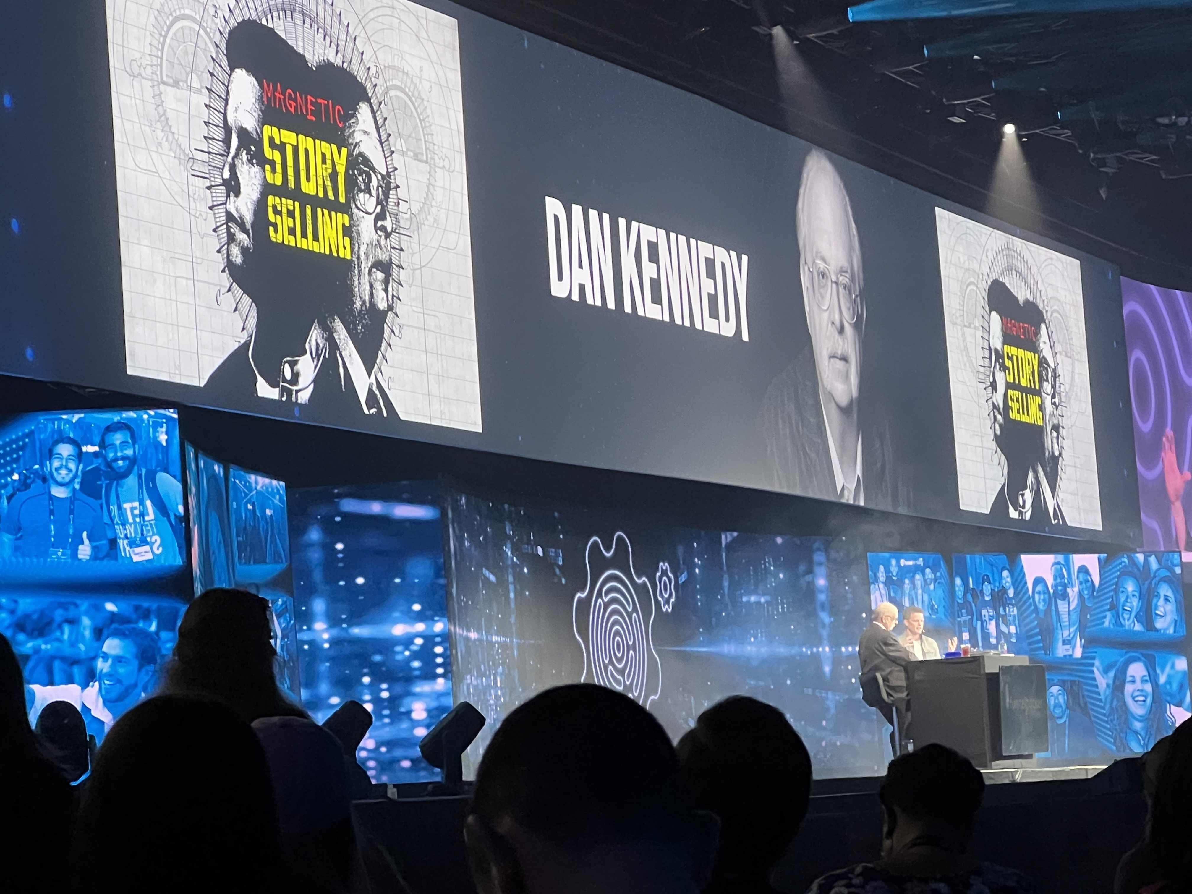 Russell Brunson interviewing Dan Kennedy on stage with a screen advertising him