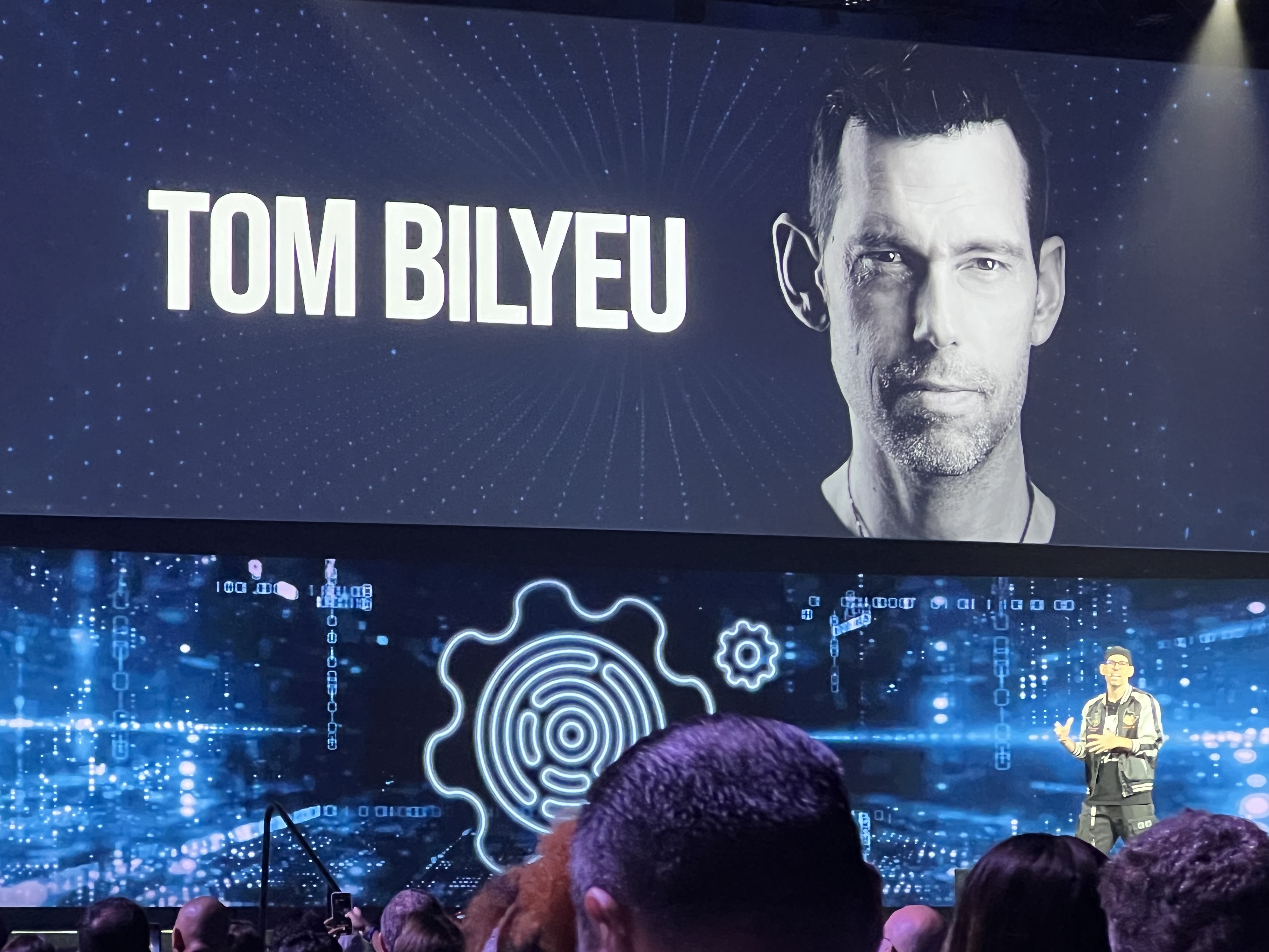 Tom Bilyeu on stage with a screen advertising him